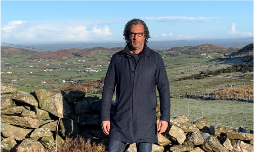 David Olusoga stood infront of a stone wall with rolling hills behind, in Ireland