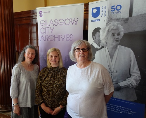 Photo of the core project team, Dr Elaine Moohan, Mrs Carol Raeside, and Dr Irene O’Brien standing in front of banners for Glasgow City Archives and The Open University Scotland 50 years (which has an image of Jenny Lee).