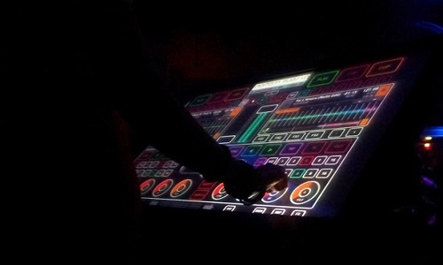 n a dark room, a digital touchscreen shines with bright colours that show a control panel in a DJ-ing software programme. Out of the darkness, a silhouetted person’s hand uses the controls.
