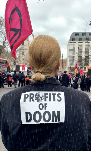 Image from Extinction Rebellion protest in London; in foreground, back of a woman wearing jacket reading Profits of Doom. 