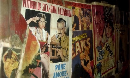 A photograph showing faded film posters that have been stuck to a wall over the top of one another, with some partially ripped away. The posters show illustrations of film personalities (including Vittoria De Sica and Gina Lollobrigida) and advertise movies such as My Fair Lady