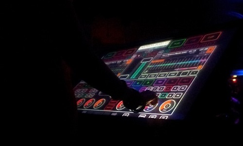 In a dark room, a digital touchscreen shines with bright colours that show a control panel in a DJ-ing software programme. Out of the darkness, a silhouetted person’s hand uses the controls. 