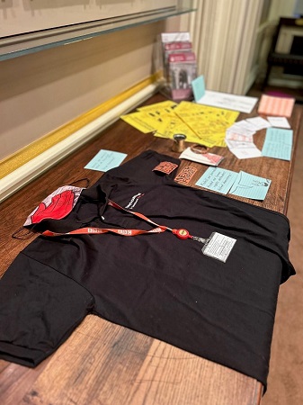 Photograph from the left side showing a display of objects related to journalism and community activism on a table. The items on the table include a black press t-shirt and a red press ID lanyard, Some yellow cards with printed instructional images, a small pink notebook, a small brass long-handled jug, red cards reading ‘never forget’ and various cards in blue and white.
