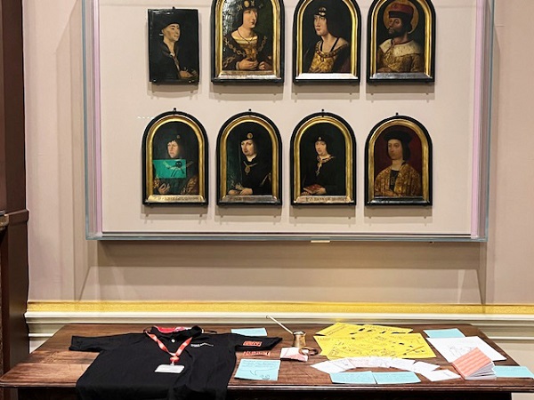 Photograph from the front showing a display of objects related to journalism and community activism on a table beneath a set of 8 Tudor portraits hung on the wall behind glass. The items on the table include a black press t-shirt and a red press ID lanyard, Some yellow cards with printed instructional images, a small pink notebook, a small brass long-handled jug and various cards in blue and white. 