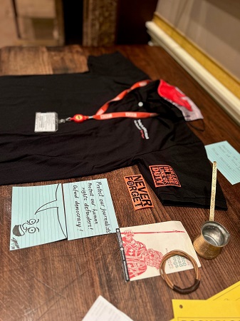 Photograph from the right showing a display of objects related to journalism and community activism on a table. The items on the table include a black press t-shirt and a red press ID lanyard, a small brass long-handled jug and woven bracelet, red cards reading ‘never forget’ and ‘killed in the line of duty’ and a pair of blue cards featuring a cartoon character and a speech bubble stating: ‘Protect our journalists! Protect our human rights defenders! Defend democracy!’