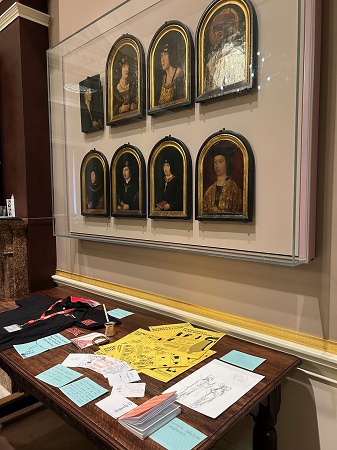 Photograph from the right showing a display of objects related to journalism and community activism on a table beneath a set of 8 Tudor portraits. At the far end of the table is a black press t-shirt and a red press ID lanyard, and at the near end is a monochrome sketch on white paper, a pink notebook and a set of yellow instructional cards.