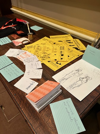 Photograph from the right showing a display of objects related to journalism and community activism with a close-up view of a monochrome sketch on white paper, which shows a figure with a loudspeaker mouth and a figure with large ears. A set of yellow instructional cards show how to make a makeshift tear-gas mask, bucket pamphlet bomb and other activism tools. A blue handwritten card reads ‘Grassroots education and materials’.