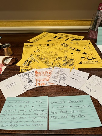 Photograph from the front showing a display of objects related to community activism with a close-up view of handwritten zines, and yellow instructional cards for activists.