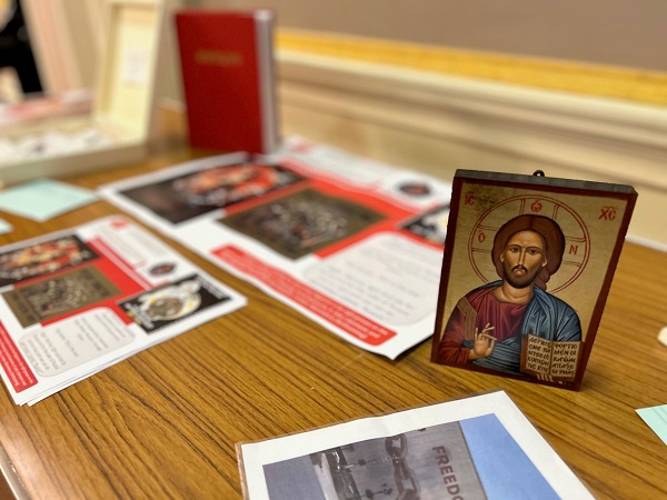 Photograph showing a close-up of an Orthodox icon. In front of it, the corner of a photograph of a wall in a public space is visible, which has been painted with the word ‘freedom’ and an image of chains. Behind them is a collection of posters featuring neo-Pagan imagery, a large red hardback book and an open square wooden box.