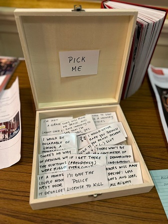 Photograph showing a square wooden book. The text in the lid reads ‘Pick me’ and the box contains a collection of badges featuring quotes from Brazil’s former President Bolsonaro. The foremost badge reads ‘I’d give the police license to kill’.