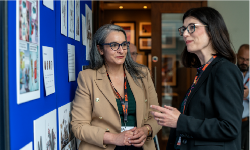 Two female academics from the organising committee stand discussing the exhibition of academic-artist collaborative cartoons at the 2024 BISA conference