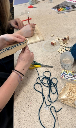 The hands of two students are visible, in front of a desk loaded with art supplies. One is cutting, newspaper fragments and the other is gluing them. 