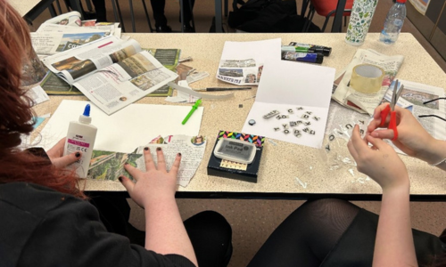 The hands of two teenagers are visible. One is sketching on a wooden plaque and the other is threading wool through cardboard.