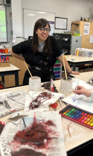 An artist in a paint-splattered apron stands smiling at the front of an art studio. She is leaning on a desk on which the work-in-progress of students is visible.