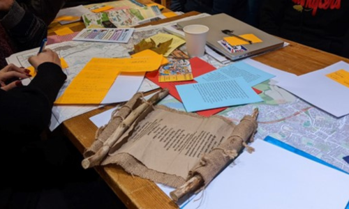 Photo taken as part of the focus group. A focus on a participate writing on a post-it note. They are surrounded by paper, maps and other collaboration tools. 