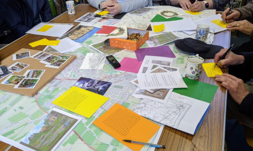 Photo taken from the project focus group. A group of people sat around a table with a scattering of maps, postcards, photos and recording devices showing active collaboration in progress