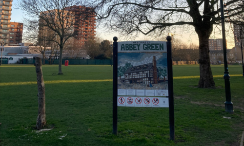 The image shows Abbey Green in the centre of Barking. At the forefront is a noticeboard sign with an image of an historic building with “Abbey Green” written above; in the background is an open green space with trees, flanked by new high-rise apartment blocks