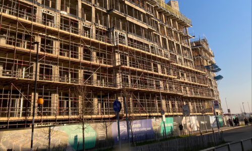 The image shows a building being constructed in Barking Riverside. The image shows the eight-story bare concrete structure, surrounded by scaffolding with a crane hovering above. Below are people waiting at a bus stop and signs on the building cladding advertising the new homes being built.
