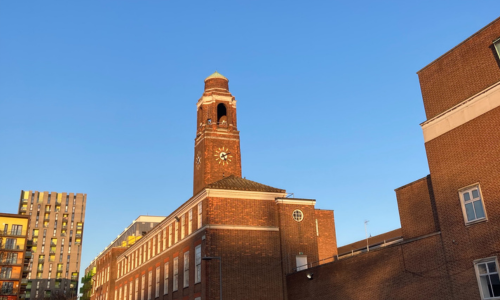 The image shows Barking Town Hall viewed from the rear, with its tall red-brick clocktower reflecting the light of sunrise. Beside it is a newly built apartment block and an empty street.