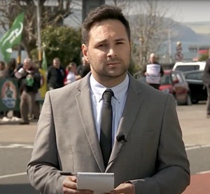 Person in a suit holding a notepad and microphone, standing outdoors with a group of people and cars in the background