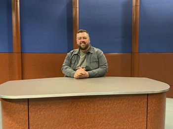 Person seated at a news desk in a studio with blue and wood-panelled background