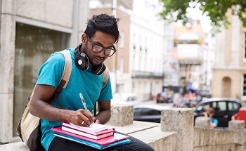 Asian young male student sitting on wall outdoors during the day writing in textbook and wearing headphones