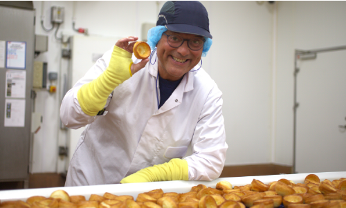Presenter Gregg Wallace, wearing protective clothing, holds up a small Yorkshire Pudding next to a conveyor belt full of Yorkshire Puddings, in a factory in Hull