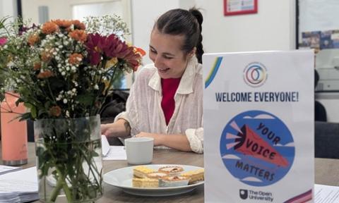 A meeting table with flowers, a plate of cakes, and a sign reading “Welcome Everyone! Your Voice Matters – The Open University,” with a person seated behind papers and a cup