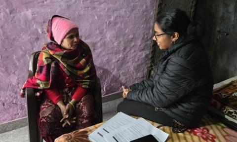 Two indian women sit indoors having a discussion, with documents on a bed and one person wrapped in patterned clothing.