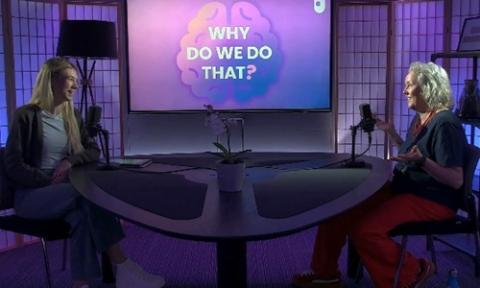 Podcast recording in a studio, with two people seated across a curved table speaking into microphones. A screen in the background displays the title “Why Do We Do That?”