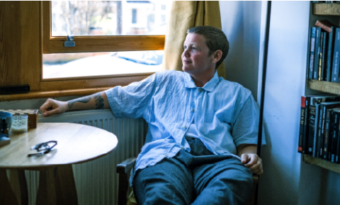 Kae Tempest sits comfortably in a chair next to a radiator between a small table and a full bookcase