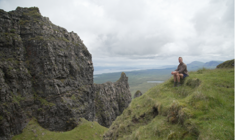 Ben Fogle sits at the top of the Quiraing on Skye with rock formations and the coast behind him in the distance