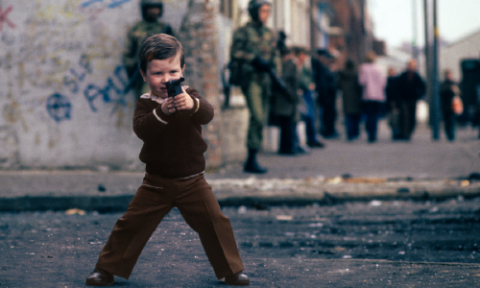 A young boy plays with a toy pistol on a street in Belfast with people and soldiers in the background