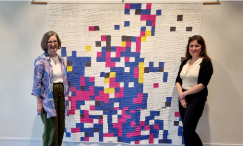 Heather Richardson and Bree Hocking stand either side of a large patchwork blanket hanging on the wall. The blanket is based on a map and is made up of white, blue, pink, yellow and black squares. 