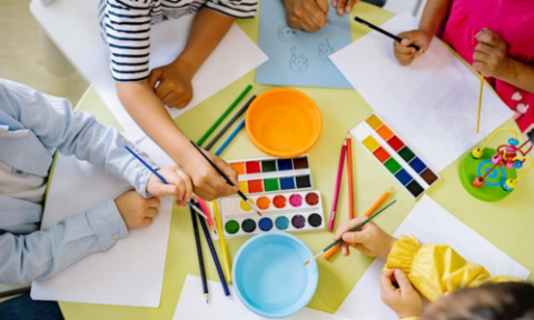 A group of five children gathered around a table, covered with water paints, paper and paintbrushes