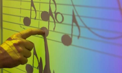 A teacher’s hand points to musical notes on a colourful interactive whiteboard
