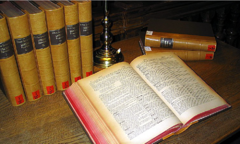 An open book on top of a wooden table with a stake of seven books behind, next to a candle