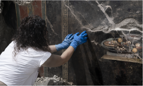 Archaeologist Roberta Frisco, wearing blue gloves, uncovers a perfectly preserved wall fresco depicting an ancestor of a pizza on a silver platter as part of a meal depicted in new excavations in Pompeii