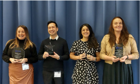 Four students stand holding their Award trophies in front of a blue background curtain, all smiling with pride